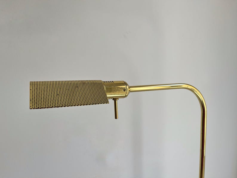 May include: A vintage brass desk lamp with a perforated shade. The lamp has a rectangular shade with a grid of small holes, a curved arm, and a vertical support. The brass finish gives the lamp a classic, retro look.