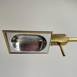 May include: A vintage brass desk lamp with a rectangular head and a long, adjustable arm. The lamp head has a perforated border and houses a lightbulb. The arm and base are a shiny gold colour.