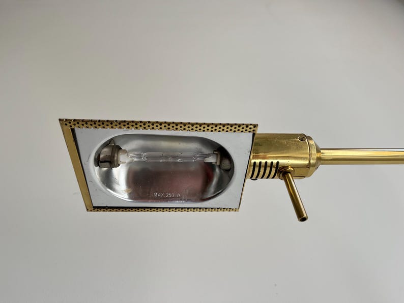May include: A vintage brass desk lamp with a rectangular head and a long, adjustable arm. The lamp head has a perforated border and houses a lightbulb. The arm and base are a shiny gold colour.
