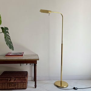 Peut inclure: Un lampadaire doré réglable avec un abat-jour rectangulaire. La lampe a un bras fin et incurvé et une base ronde. Un accent en bois brun est visible sur le poteau de la lampe. La lampe est branchée sur une prise murale. Une petite table et un panier en osier sont également dans l'image.