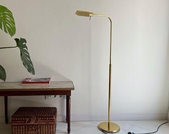 May include: A gold-toned, adjustable floor lamp with a rectangular shade. The lamp has a slender, curved arm and a round base. A brown wooden accent is visible on the lamp's pole. The lamp is plugged into a wall outlet. A small table and a woven basket are also in the image.