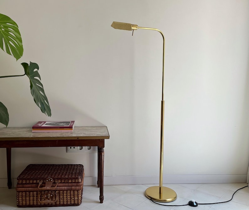 May include: A gold-coloured, adjustable floor lamp with a rectangular shade. The lamp has a slender, curved arm and a round base. A brown wooden accent is visible on the lamp's pole. The lamp is plugged into a wall outlet. A small table and a woven basket are also in the image.