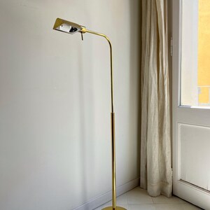 May include: A gold-coloured, vintage floor lamp. It has a rectangular light fitting, a curved arm, and a tall, slender pole. The base is a circular disc. The lamp is positioned in a corner next to a window with a beige curtain.