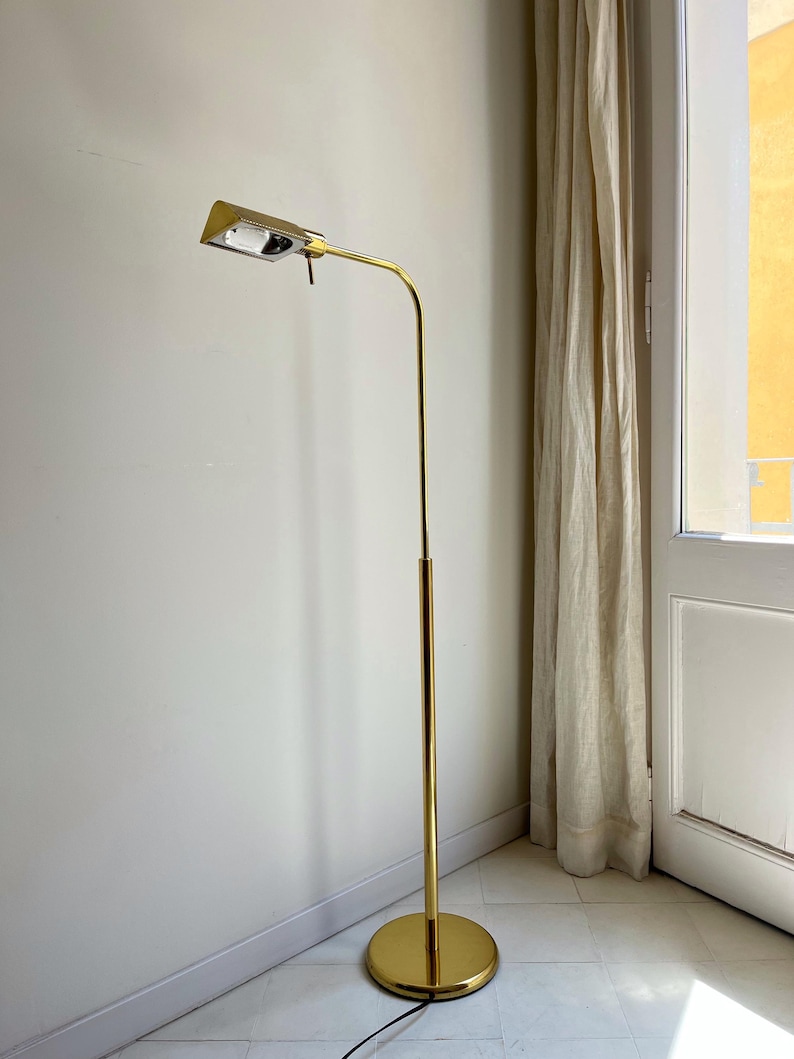 May include: A gold-coloured, vintage floor lamp. It has a rectangular light fitting, a curved arm, and a tall, slender pole. The base is a circular disc. The lamp is positioned in a corner next to a window with a beige curtain.