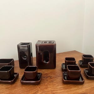 Könnte beinhalten: Eine Sammlung dunkelbrauner Keramik-Geschirr, darunter quadratische Tassen, Untertassen und zwei rechteckige Vasen. Das Set ist auf einer Holzoberfläche angeordnet und zeigt ein minimalistisches Design.