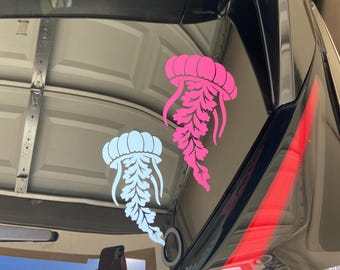 May include: Two vinyl decals applied to a car window, featuring a bright pink jellyfish positioned above a light blue jellyfish. Each jellyfish design displays a rounded bell top and long, intricate, leaf-like trailing tentacles.