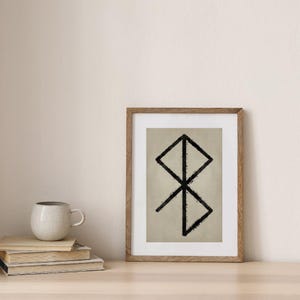 Könnte beinhalten: Ein gerahmter Druck mit einem schwarzen geometrischen Design auf beigem Hintergrund. Das Kunstwerk befindet sich in einem Holzrahmen und ruht auf einer hellen Oberfläche. Ein Stapel Bücher und eine gesprenkelte Tasse sind ebenfalls zu sehen.