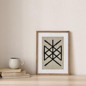 May include: A framed art print featuring a black geometric design on a beige background. The artwork is in a wooden frame and rests on a wooden surface. A stack of books and a ceramic mug are also on the surface.