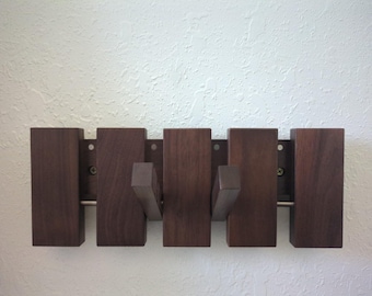 May include: A wooden wall-mounted coat rack with five dark brown rectangular blocks. Each block has a hook extending from it. The rack is mounted on a light-colored wall, providing a minimalist and functional design.