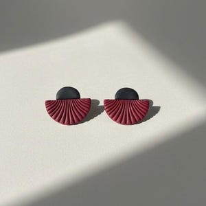Pode incluir: Um par de brincos de pressão feitos à mão. Cada brinco apresenta um design em forma de leque na cor vermelho escuro, com um topo preto semicircular. Os brincos são exibidos em uma superfície branca.