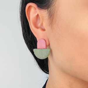 Op de afbeelding: Een close-up van een roze en groene oorbel. De oorbel heeft een roze, boogvormige bovenkant en een halfronde basis met verticale groene lijnen. De oorbel is bevestigd aan een oor.