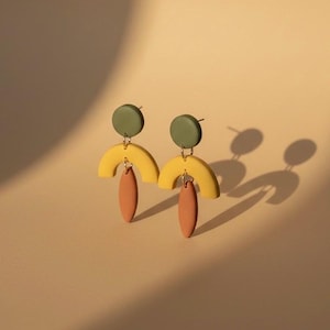May include: A pair of abstract earrings. Each earring has a small olive green circle, a yellow arch, and a peach-coloured teardrop shape. The earrings are displayed on a beige background.