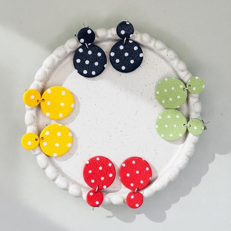 May include: A collection of colorful polka dot earrings in black, yellow, red, and green. Each pair features a circular design with white dots. The earrings are displayed on a white, scalloped-edge plate.