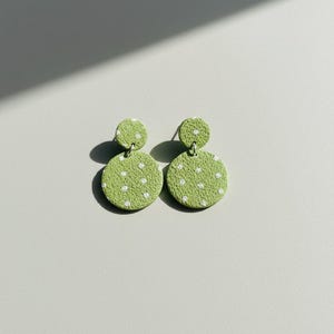 May include: A pair of green, polka-dot earrings. Each earring features a small, round stud connected to a larger, circular drop. The earrings are a light green color with white polka dots.