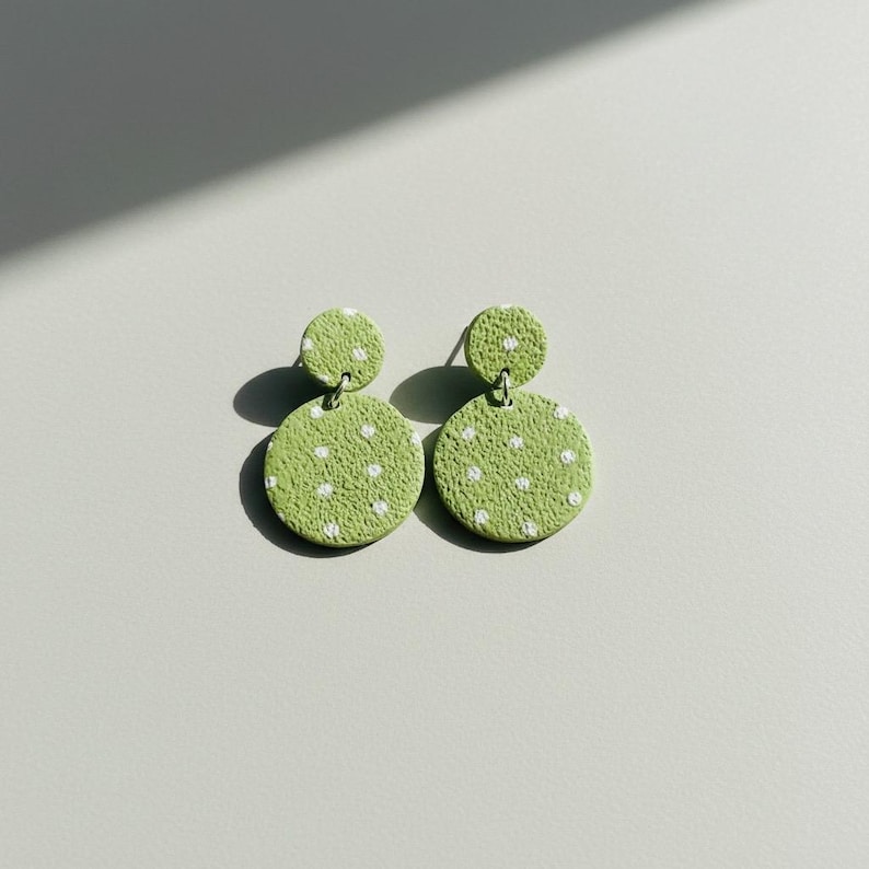 May include: A pair of green, polka-dot earrings. Each earring features a small, round stud connected to a larger, circular drop. The earrings are a light green color with white polka dots.