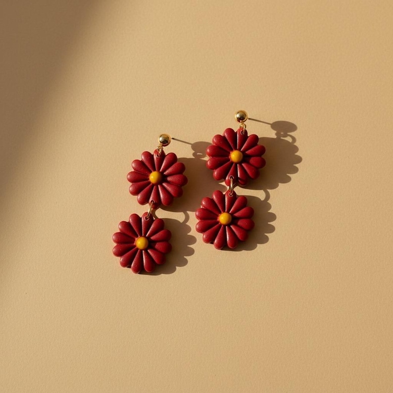 May include: A pair of daisy earrings. Each earring features two burgundy daisy flowers with yellow centers, suspended from a gold-colored stud. The earrings are set against a beige background, with shadows adding depth.