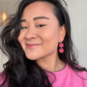 May include: A close-up shot of a person wearing pink daisy earrings. The earrings are composed of two stacked, bright pink flowers with yellow centers. The person is wearing a pink shirt and has long, dark hair.