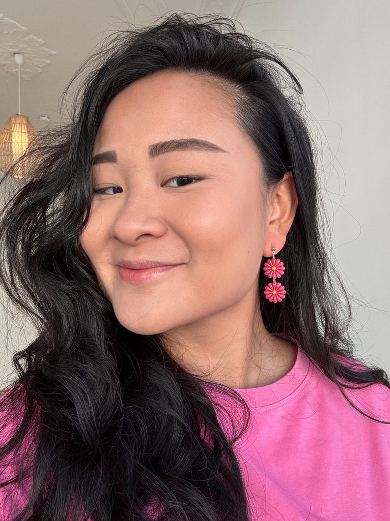 May include: A close-up shot of a person wearing pink daisy earrings. The earrings are composed of two stacked, bright pink flowers with yellow centers. The person is wearing a pink shirt and has long, dark hair.