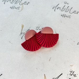 以下が含まれることがあります： 赤とピンクの粘土で作られた手作りのイヤリングのペア。 イヤリングは、上部にピンクの半円と、テクスチャ加工された赤い扇形の底を備えた扇のようなデザインが特徴です。 イヤリングは、テクスチャ加工されたオフホワイトの表面にあります。