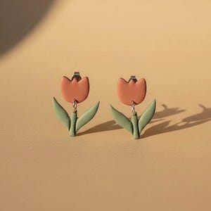 Könnte beinhalten: Ein Paar Ohrringe in Tulpenform. Jeder Ohrring hat einen pfirsichfarbenen Blütenkopf und zwei grüne Blätter. Die Ohrringe hängen an silberfarbenen Ohrhaken, vor einem neutralen Hintergrund.