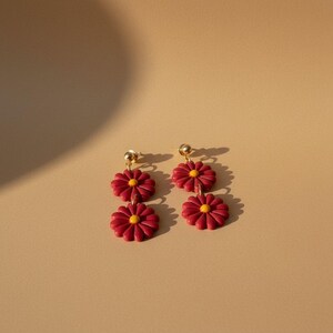 May include: A pair of dangle earrings featuring a daisy design. Each earring has two red daisy flowers with yellow centers and gold-colored posts. The earrings are set against a neutral background.