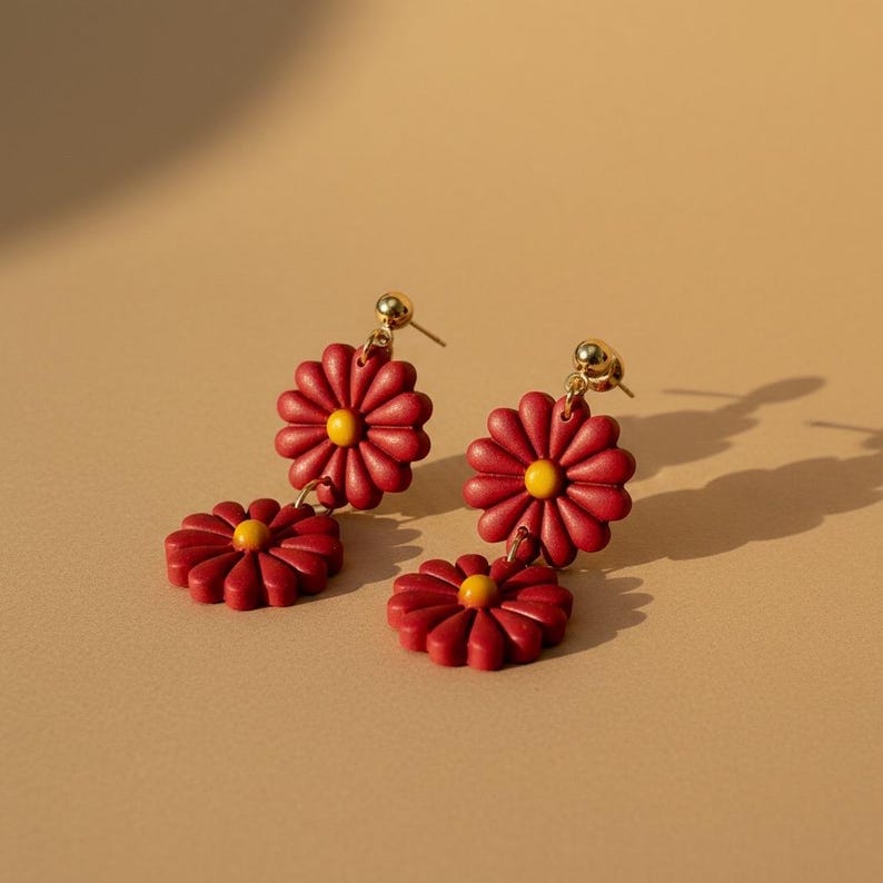 May include: A pair of red daisy earrings with gold-colored posts and yellow centers. Each earring features two daisy flower pendants. The earrings are set against a neutral background.
