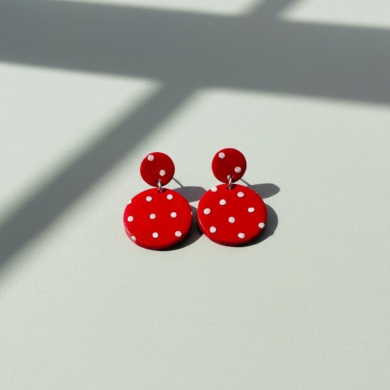 May include: A pair of red polka dot earrings. Each earring features a small, round stud connected to a larger, circular pendant. The pendants are bright red with evenly spaced white dots, creating a playful and eye-catching design. The earrings are set against a light background.