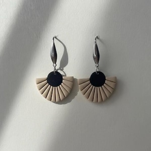 以下が含まれることがあります： 扇形のデザインが特徴的な、手作りのイヤリングのペア。イヤリングは、黒い円形の中心部と、その下にベージュの扇形をしています。イヤリングは銀色のフックから吊り下げられています。