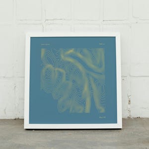 May include: A framed print featuring a topographic map design. The artwork has a blue background with gold contour lines. The text "Cairn Gorm" and "1245 m" are at the top, and "Blue Hill" is at the bottom. The frame is white.