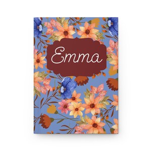 May include: A rectangular notebook with a periwinkle blue background featuring a pattern of orange, yellow, and blue flowers with brown stems. A dark red scalloped label is centered on the cover with the name Emma written in white script.
