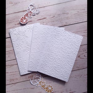 Könnte beinhalten: Weiße geprägte Grußkarten mit Schneeflocken- und Blumenmustern. Zwei dekorative Hirsch-Ausschnitte mit braunen und goldenen geometrischen Designs sind ebenfalls vorhanden. Die Karten liegen auf einer hellen Holzmaserung.