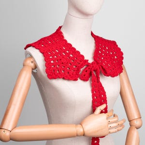 May include: A bright red, crocheted collar with a scalloped edge and a tie closure. The collar is displayed on a mannequin, showcasing its intricate openwork design. This handmade accessory adds a touch of vintage charm.