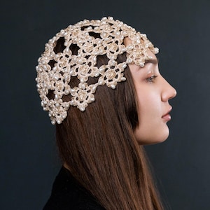 Könnte beinhalten: Ein cremefarbenes Häkel-Kopfstück mit floralem Design, verziert mit kleinen, runden Perlen. Das Kopfstück wird getragen, mit langen, braunen Haaren, die nach hinten fallen. Der Hintergrund ist eine dunkle, einfarbige Farbe.