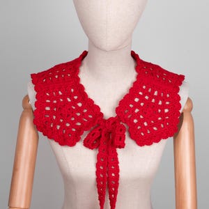 May include: A bright red, crocheted collar with a scalloped edge and a tie closure. The collar is displayed on a mannequin, showcasing its openwork design and the delicate texture of the yarn. The collar is a fashion accessory.