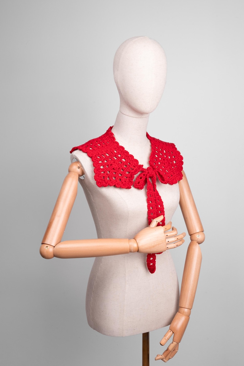 May include: A bright red, crocheted collar with a scalloped edge and a tie closure. The collar is displayed on a mannequin, showcasing its intricate openwork design. This handmade accessory adds a touch of vintage charm.