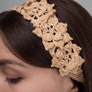 Könnte beinhalten: Ein hellbraunes Häkelstirnband mit floralem Design. Das Stirnband besteht aus einem gewebten Material und wird über der Stirn getragen. Das Blumenmuster besteht aus mehreren Blütenformen.