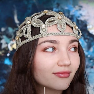 K&ouml;nnte beinhalten: Ein goldfarbenes, geh&auml;keltes Diadem mit floralen und Schmuckakzenten. Der Kopfschmuck wird auf dem Kopf einer Person getragen, im Hintergrund befindet sich ein blau-wei&szlig;es abstraktes Design. Das Diadem ist ein dekoratives Accessoire.