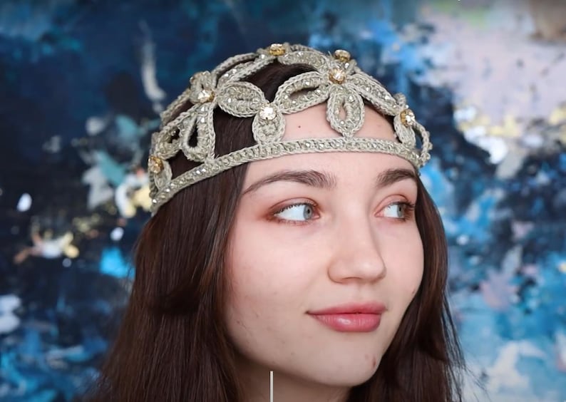 K&ouml;nnte beinhalten: Ein goldfarbenes, geh&auml;keltes Diadem mit floralen und Schmuckakzenten. Der Kopfschmuck wird auf dem Kopf einer Person getragen, im Hintergrund befindet sich ein blau-wei&szlig;es abstraktes Design. Das Diadem ist ein dekoratives Accessoire.