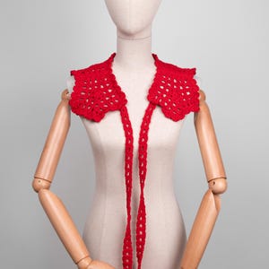 May include: A bright red, crocheted collar with a scalloped edge and long, dangling ties. The openwork design is displayed on a mannequin, showcasing the intricate details of the handmade accessory. This collar adds a pop of color and texture.