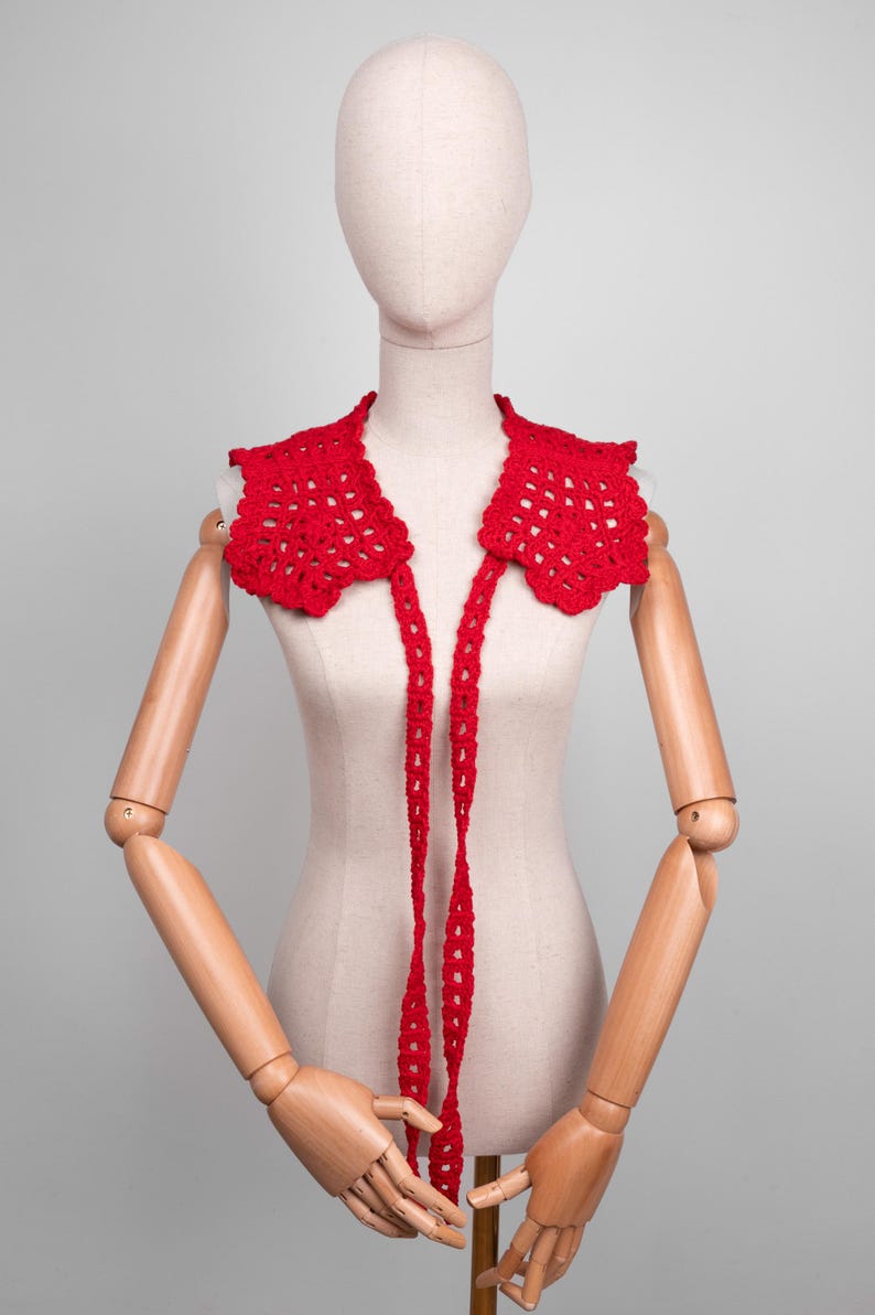 May include: A bright red, crocheted collar with a scalloped edge and long, dangling ties. The openwork design is displayed on a mannequin, showcasing the intricate details of the handmade accessory. This collar adds a pop of color and texture.