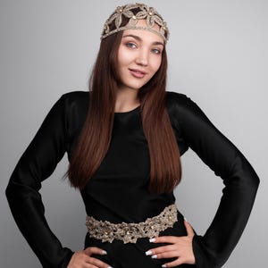 K&ouml;nnte beinhalten: Eine Frau in einem schwarzen Kleid, mit einem dekorativen goldfarbenen G&uuml;rtel und einem passenden Kopfschmuck. Der Kopfschmuck und der G&uuml;rtel sind mit floralen und Kristallverzierungen versehen. Die Frau hat lange braune Haare und l&auml;chelt.