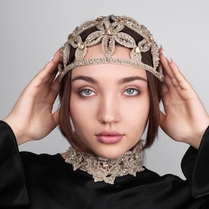 K&ouml;nnte beinhalten: Ein goldfarbenes, geh&auml;keltes Kopfst&uuml;ck und eine passende Halskette. Das Kopfst&uuml;ck hat die Form einer Krone mit floralen Designs und ist mit kleinen, funkelnden Kristallen verziert. Das Halsband hat einen gewellten Rand und ebenfalls Kristalle. Die Frau tr&auml;gt ein schwarzes Oberteil.
