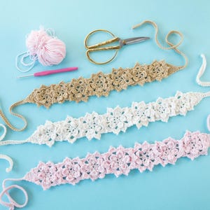 Puede incluir: Tres gargantillas de ganchillo en beige, blanco y rosa, cada una con un diseño floral. Una madeja de hilo rosa, un ganchillo y unas tijeras doradas también están sobre una superficie azul claro. Las gargantillas tienen lazos largos.