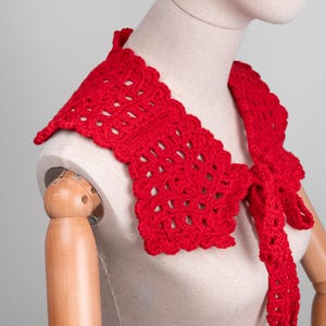 May include: A bright red, crocheted collar with a scalloped edge and a tie closure. The openwork design of the collar is visible, and it is displayed on a mannequin. The collar is a vibrant accessory.