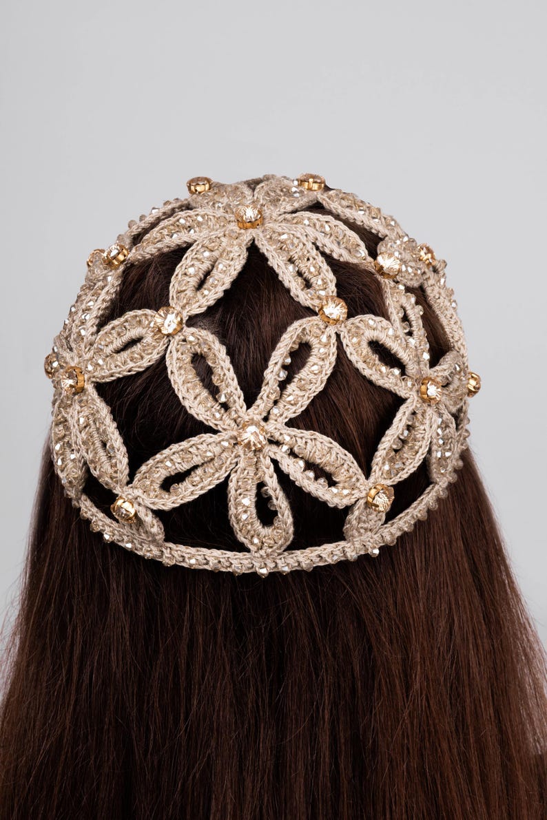 K&ouml;nnte beinhalten: Ein Kopfschmuck mit floralem Design. Der Kopfschmuck besteht aus einem hellbeigen geh&auml;kelten Material und ist mit glitzernden, goldfarbenen Verzierungen versehen. Er wird auf braunem Haar getragen.