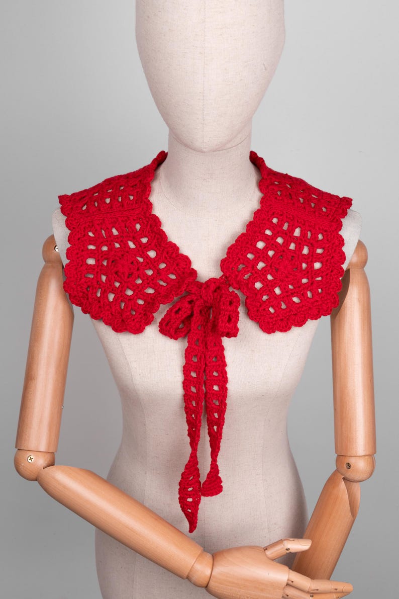 May include: A bright red, crocheted collar with a scalloped edge and a tie closure. The collar is displayed on a mannequin, showcasing its openwork design and the delicate texture of the yarn. The collar is a fashion accessory.