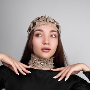 K&ouml;nnte beinhalten: Ein goldfarbenes, perlenbesetztes Stirnband und eine passende Choker-Halskette. Der Schmuck weist komplizierte Blumen- und Blattmuster mit funkelnden Akzenten auf. Die Frau tr&auml;gt ein schwarzes Oberteil vor grauem Hintergrund.
