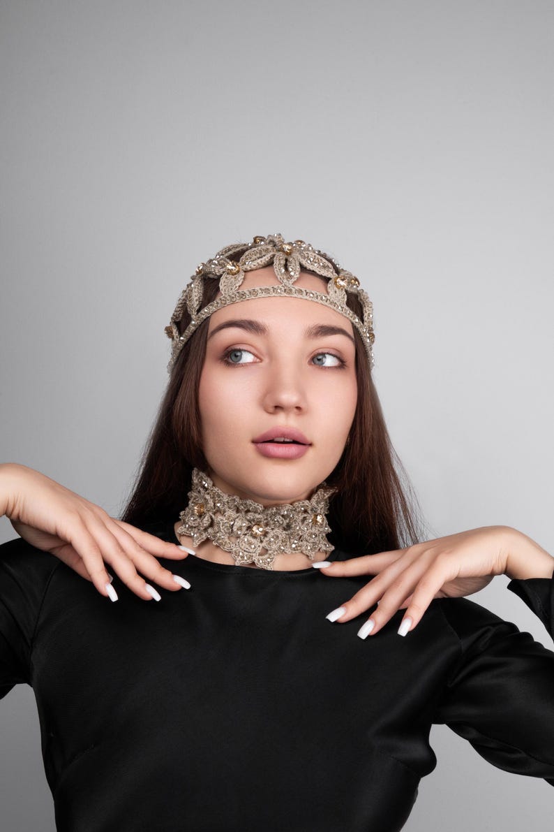 K&ouml;nnte beinhalten: Ein goldfarbenes, perlenbesetztes Stirnband und eine passende Choker-Halskette. Der Schmuck weist komplizierte Blumen- und Blattmuster mit funkelnden Akzenten auf. Die Frau tr&auml;gt ein schwarzes Oberteil vor grauem Hintergrund.