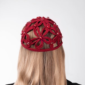 Puede incluir: Un tocado de ganchillo burdeos con un diseño floral, adornado con pequeñas cuentas rojas. El tocado se lleva puesto, con pelo largo y rubio visible. El fondo es blanco liso.
