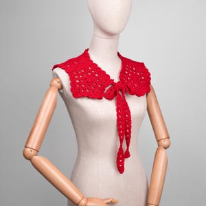 May include: A bright red, crocheted collar with a scalloped edge and long, tie-up closures. The collar is displayed on a mannequin with wooden arms and hands, against a neutral gray background. The collar is a handmade accessory.